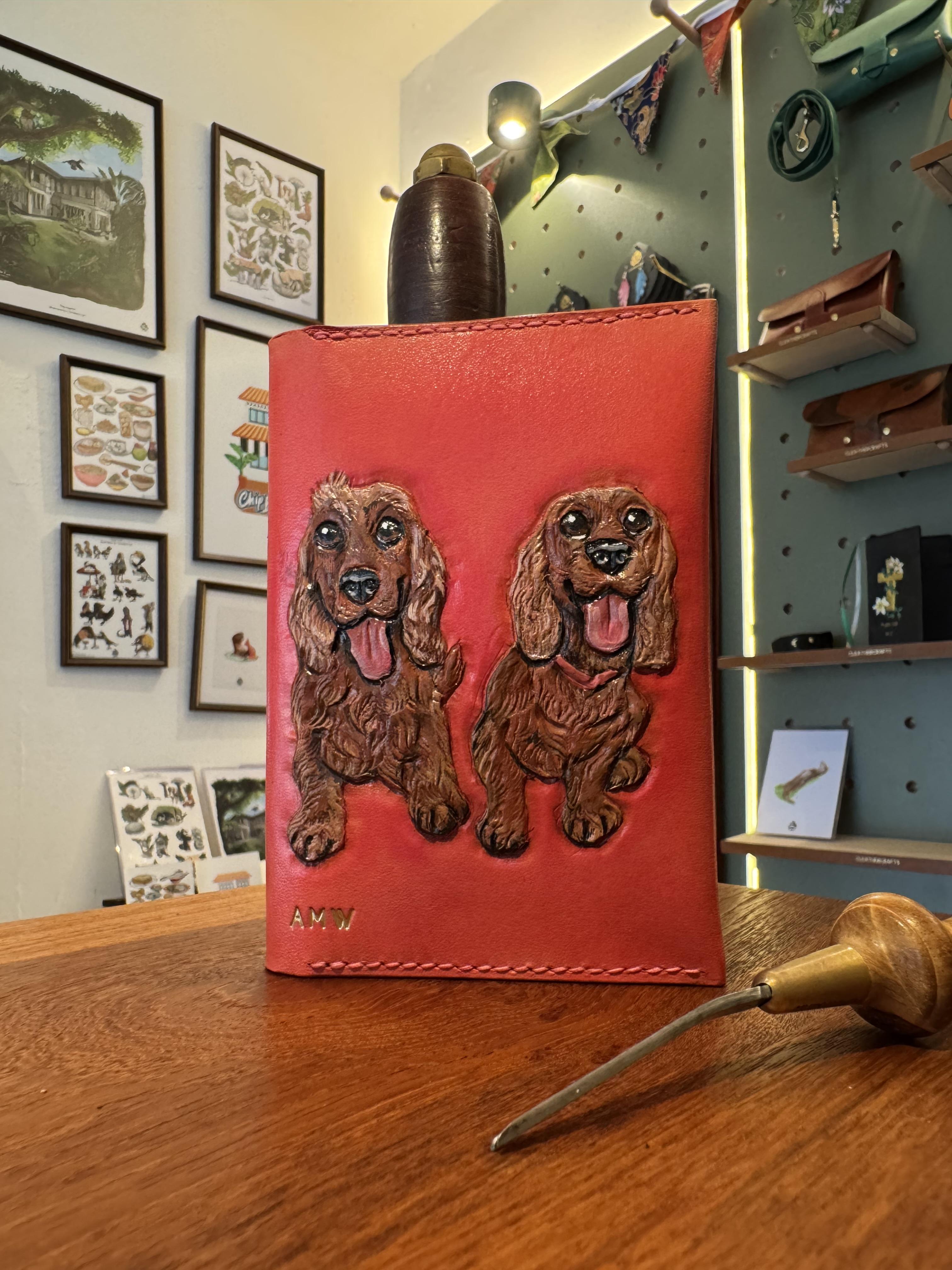 A passport holder featuring two carved Cocker Spaniels of a loving owner. Made as a gift, this was custom dyed with a pink background with the pups' names gold foiled on the interior side.